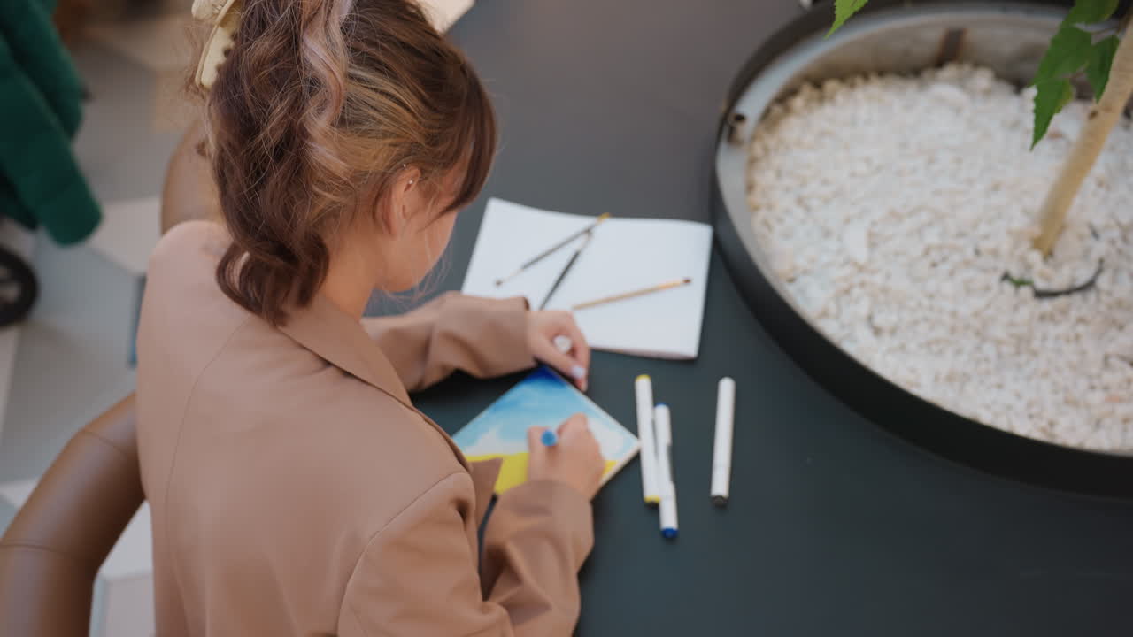 Mujer creando coloridas obras de arte en un espacio abierto, joven artista trabajando cuidadosamente en ilustraciones detalladas a color, adolescente dedicada al dibujo y coloreado precisos en una mesa redonda en un atrio