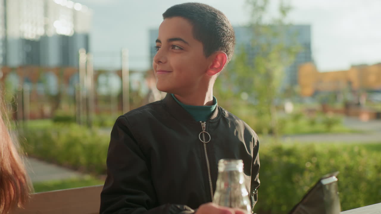 Smiling kid collects lunch from elder sister and drinks happily while sitting outdoors, with blurred view of people passing by in background