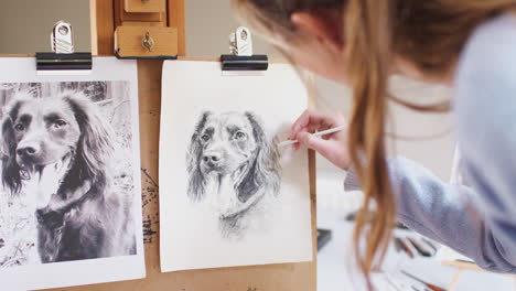 Female-Teenage-Artist-Sitting-At-Easel-Drawing-Picture-Of-Dog-From-Photograph-In-Charcoal