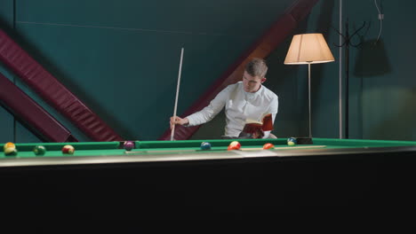 billiard player in white shirt sits on white chair reading red book while holding cue stick near green pool table with scattered billiard balls. warm lighting from floor lamp