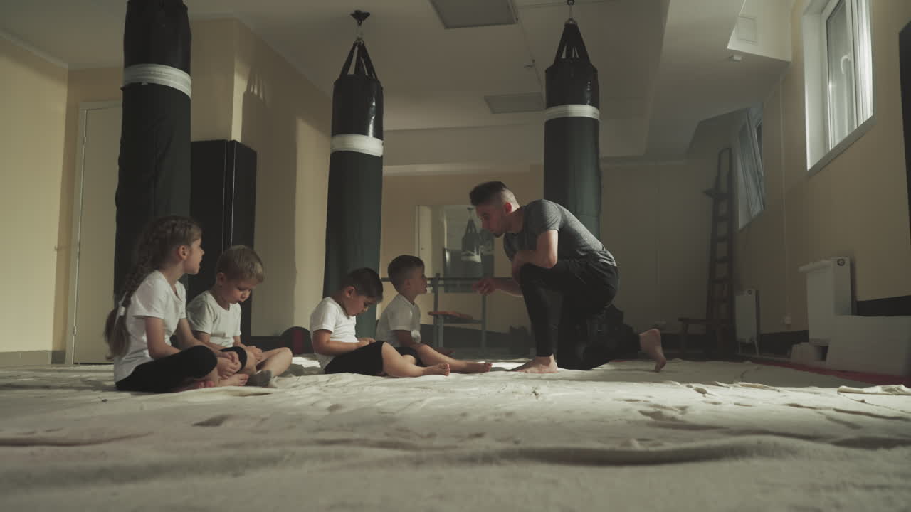 권투 클럽에서 조롱하는 강사, 지친 아이들이 바닥에 앉아서 체육관에서 전투 강사를 듣고, 무술 수업