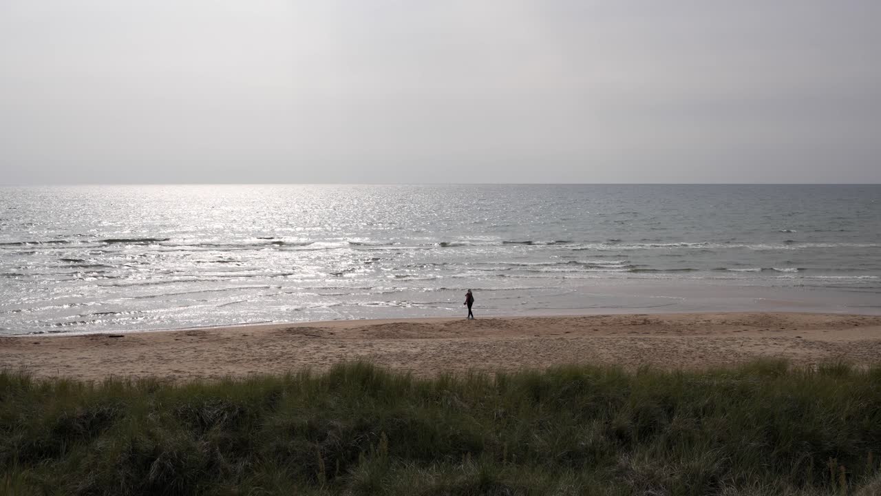 Slow-motion silhouette of a person walking along a Swedish beach. The overcast sky is pierced by a strong, diffused white light as the sun pushes through, casting an ethereal glow.