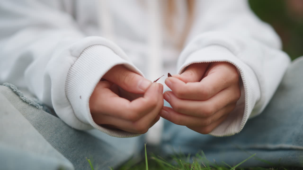 Hands gently touch grassy outdoor scene. Delicate hand movements amidst nature during serene outdoor moment. Fingers softly fidget with grass and fabric in tranquil outdoor meadow environment
