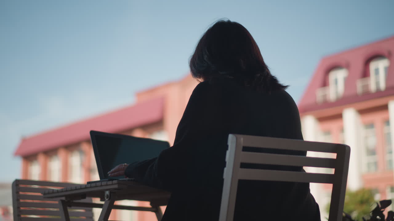 야외 도시 광장에서 나무 의자에 앉아있는 여성, 테이블 위에 스마트폰과 함께 노트북에서 일하고, 빨간 건물과 파란 하늘의 흐릿한 배경에 설정