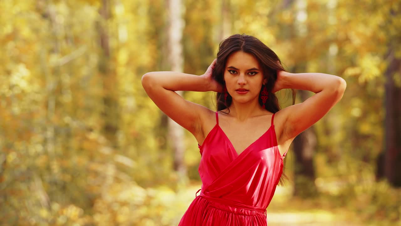A Young Woman in a Stunning Red Dress Poses Gracefully in a Vibrant Autumn Forest, Exhibiting Beauty and Elegance Amidst Nature's Golden Surroundings
