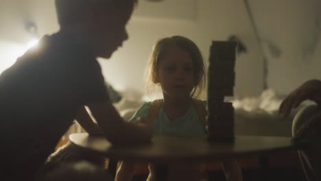 little boy gets up from table to pull block out of tower and accidentally loosens belfry. girl in background begins to worry about game. children playing board game