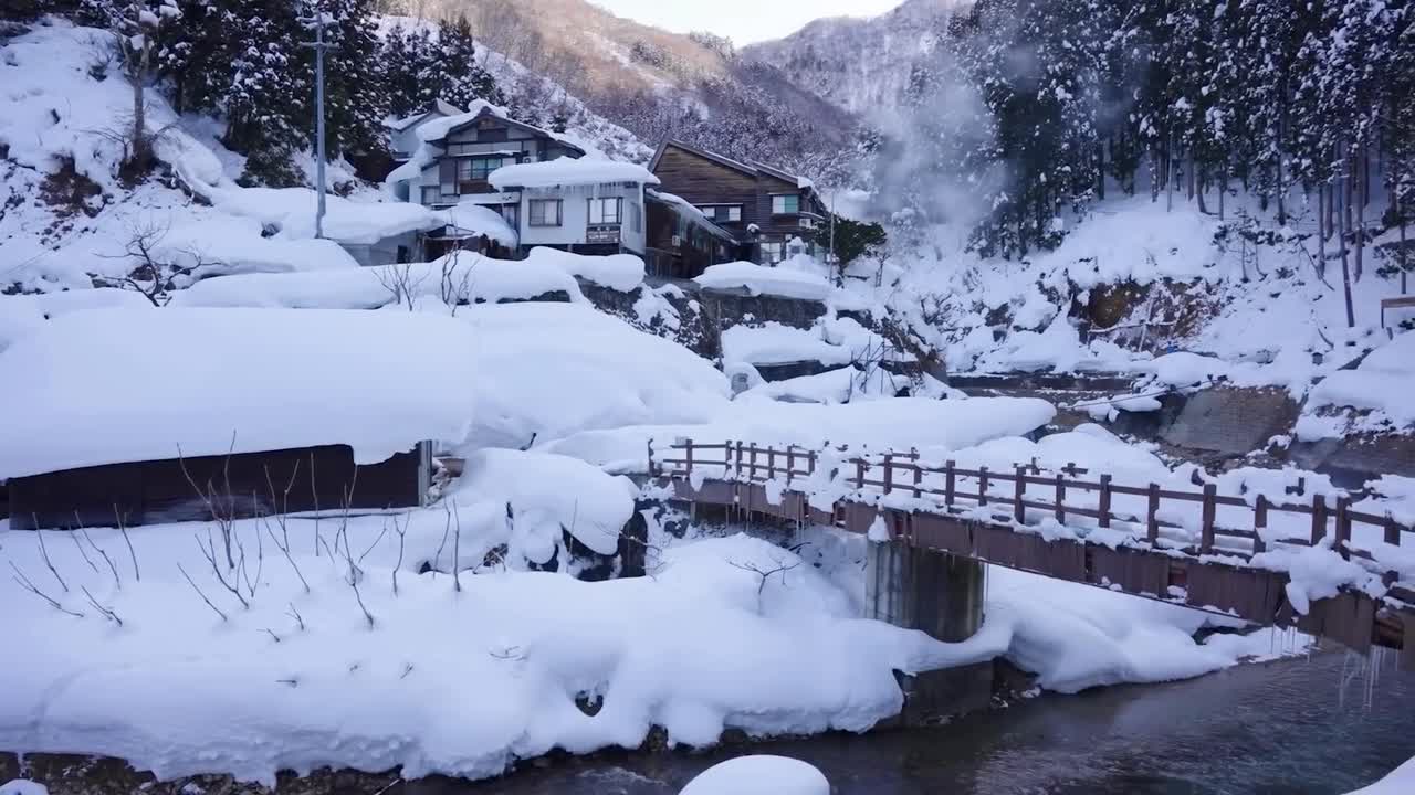 Panning footage valley with stream and snow covered landscape in Jigokudani Monkey Park, Japan. Bridge over winding flowing river with rising steam cloud. Snowy Japanese mountains in the background