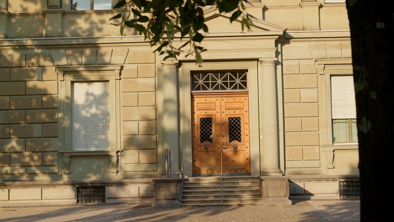L'immagine mostra l'ingresso di un edificio con doppie porte in legno e scalinata in pietra. Ombre di foglie e rami d'albero si proiettano sulla facciata