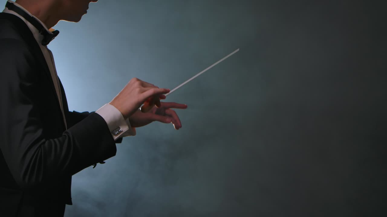 극장에서 음악가들을 동기화하고 지휘하기 위해 지휘 지이를 우아하게 흔들고 있는 사람의 측면 시각. 흑색 배경에 연기와 백라이트가 있는 옷을 입은 심포니 오케스트라 지휘자.