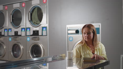 laundry owner walks toward table holding cup and notebook, placing them down while monitoring washing machines in laundromat, stainless drums spinning with clothes inside, modern equipment