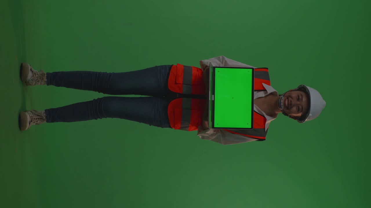 cuerpo lleno de ingeniera asiática con casco de seguridad sonriendo y mostrando portátil de pantalla verde a la cámara mientras está de pie en el estudio de fondo de pantalla verde, durante la puesta o salida del sol