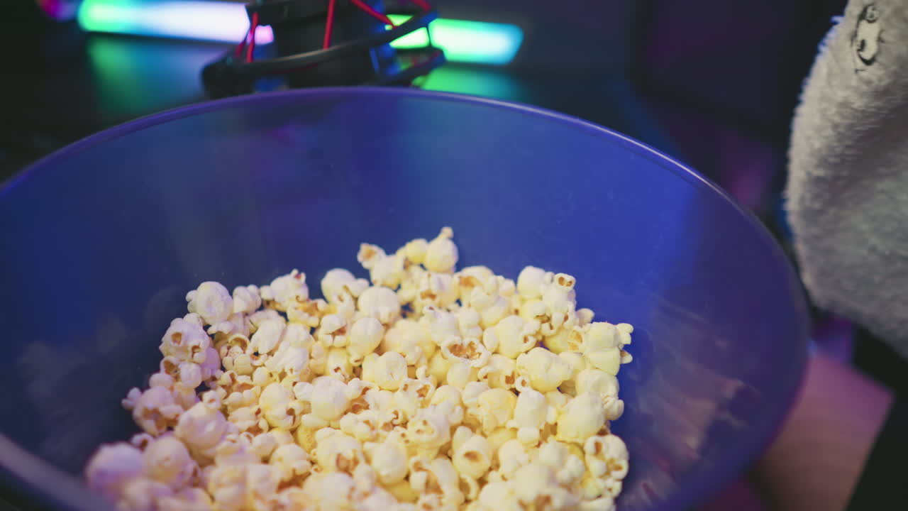 Close up of popcorn in blue bowl with person beside it, colorful blinking light reflecting off surfaces, condenser microphone partially visible above bowl