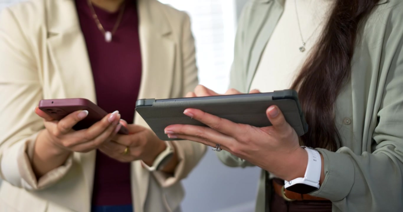 Right woman tilting tablet and tapping, starting review by window while left woman scrolling phone