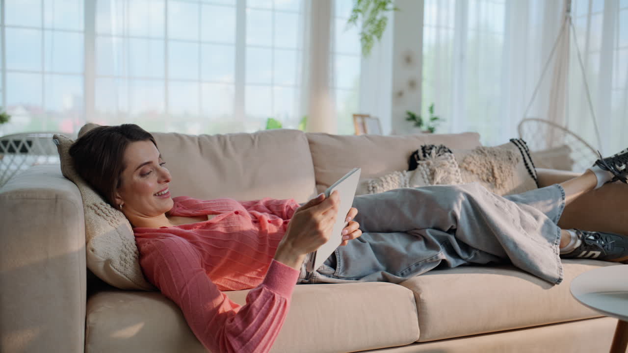 Donna che guarda un tablet sdraiata su un comodo divano. Signora sorridente che cerca contenuti