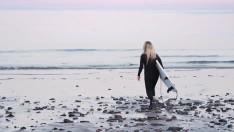 Rear-View-Of-Woman-Wearing-Wetsuit-Carrying-Surfboard-Walking-Into--Sea