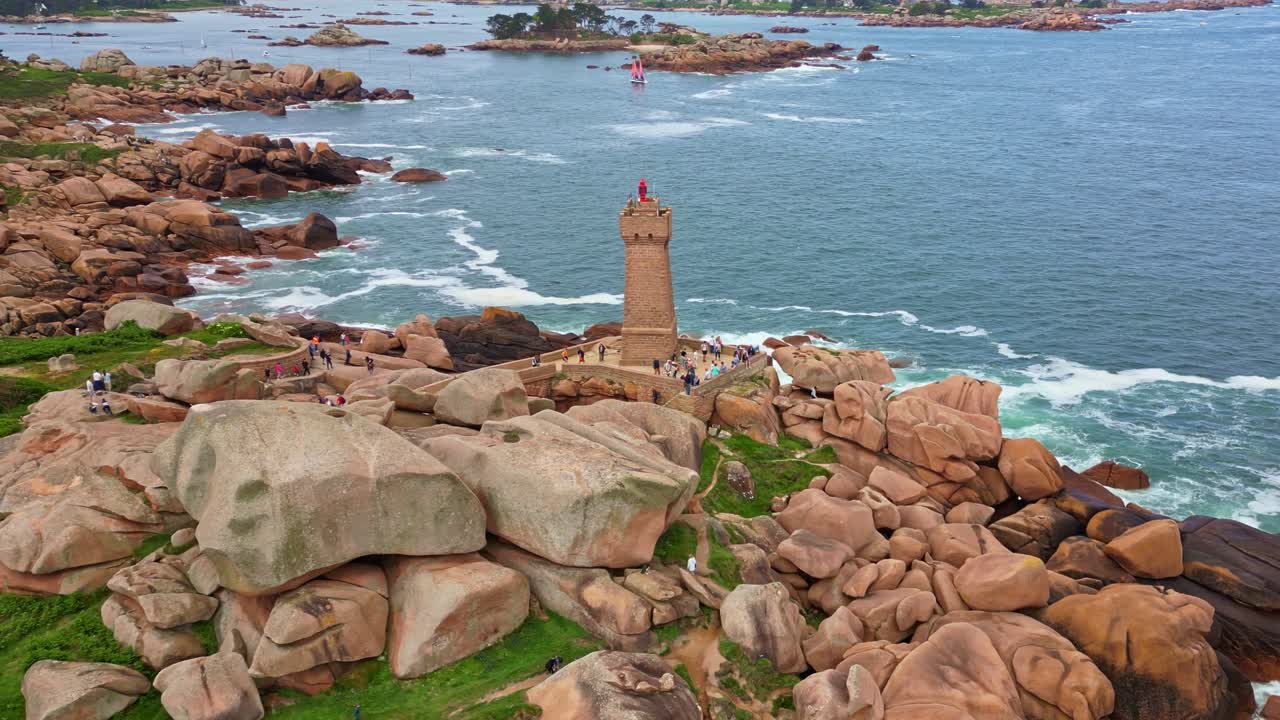 Lateral drone shot of Mean Ruz Lighthouse in Ploumanac’h, showing pink granite cliffs, crashing waves, and scattered coastal rocks along Brittany’s shoreline - France