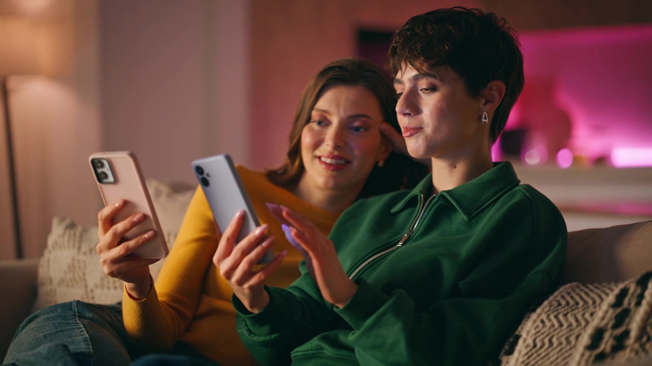 Amigas fazendo compras usando celulares, relaxando juntas no sofá aconchegante de casa, em primeiro plano