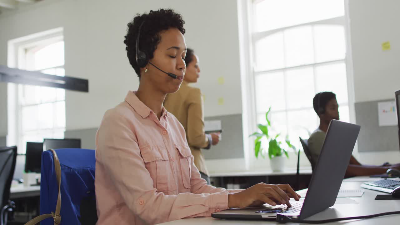 Glad afroamerikansk forretningskvinne sitter ved skrivebordet, bruker datamaskin og snakker i headset.