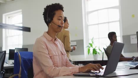 feliz mujer de negocios afroamericana sentada en un escritorio usando una computadora y hablando con auriculares