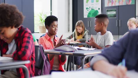 diverse female teacher and happy schoolchildren at desks learning sign language in school classroom
