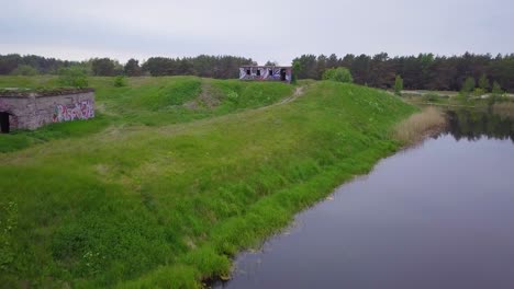 Aerial-view-of-abandoned-concrete-seaside-fortification-building,-Southern-Forts-near-the-beach-of-Baltic-sea-in-Liepaja-at-overcast-summer-day,-wide-ascending-drone-shot-moving-forward-over-the-water