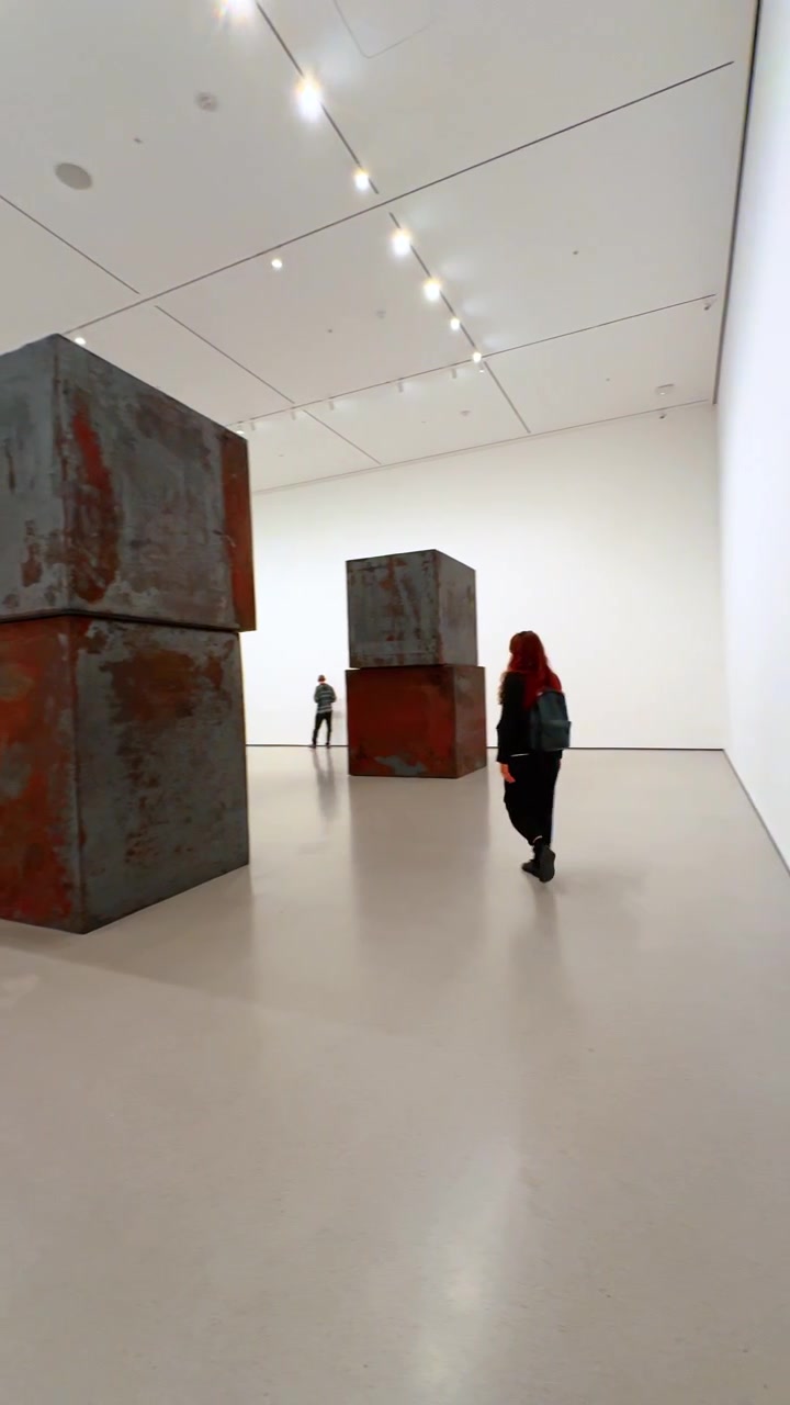 New York, USA, 12. August 2025: Besucher geht an riesiger Stahlblock-Kunst vorbei. Große patinierte Würfel schaffen eine minimalistische Szene in einem geräumigen Kunst-Innenraum