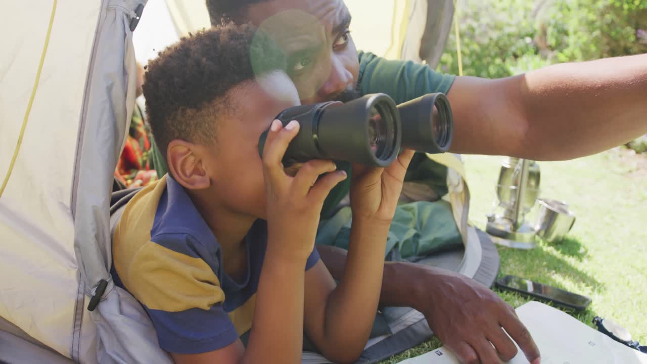 Happy african american father and son sitting in tent and looking through binoculars, in slow motion