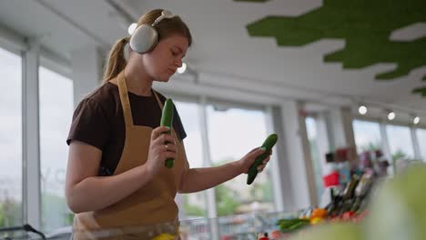 Confident-girl-in-a-brown-T-shirt-apron-and-headphones-arranges-vegetables-on-the-counter-and-listens-to-music-while-working-in-a-supermarket