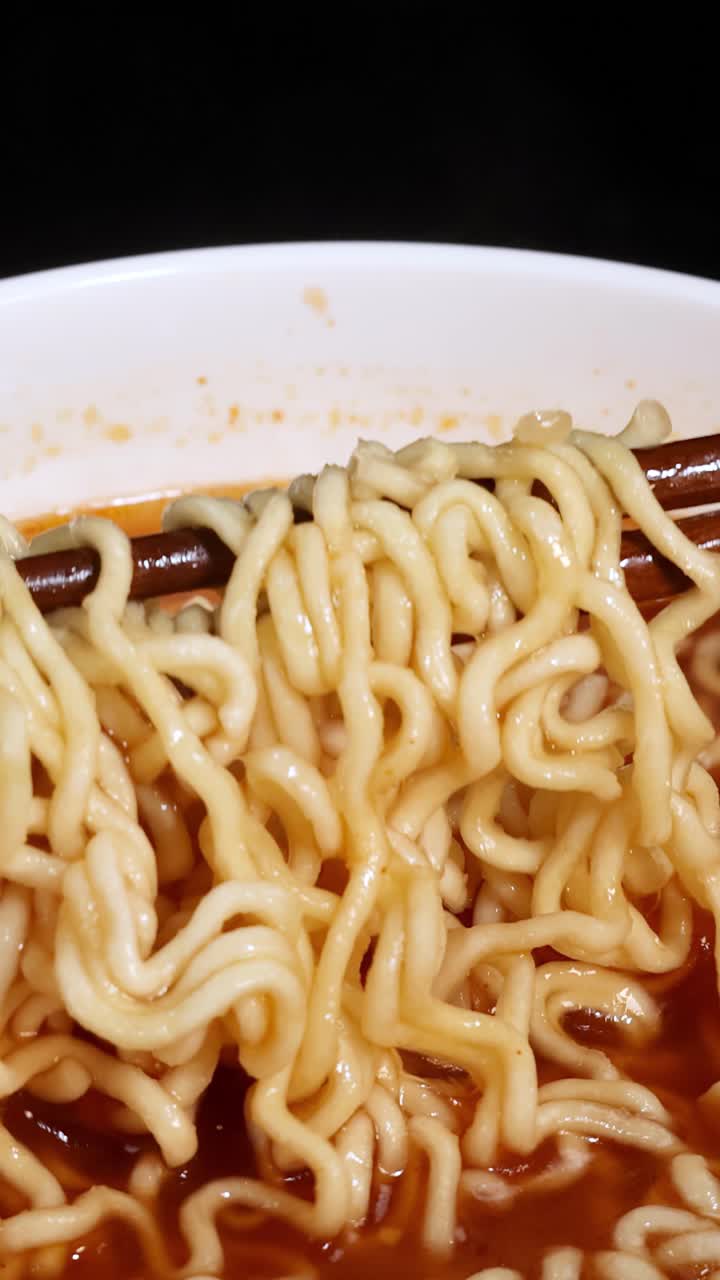 明るく均一な照明の下、クローズアップで、湯気の立つ赤いスープの丼から箸が調理されたインスタント麺を掴んで持ち上げている。