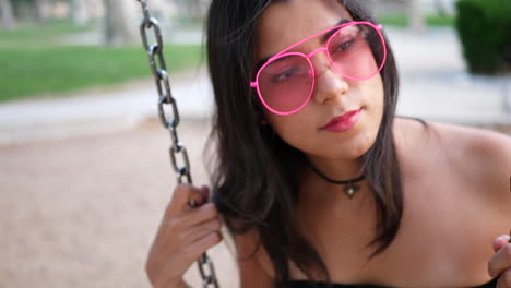 Una-Hermosa-Modelo-De-Moda-Hipster-Con-Ropa-Vintage-Y-Gafas-De-Sol-Rosas-Retro-Que-Se-Balancean-En-Un-Patio-De-Recreo-Y-Se-Divierten-A-Cámara-Lenta