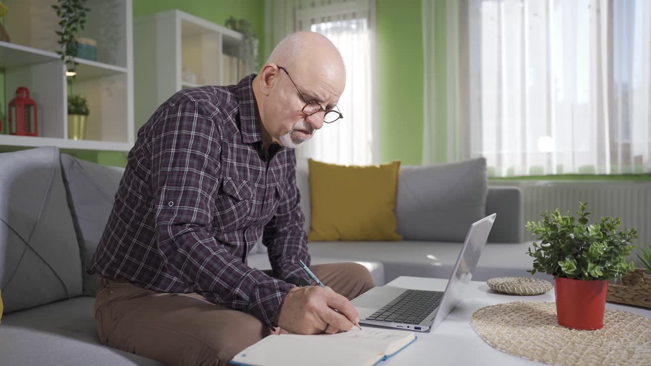 Old man using laptop at home taking notes.