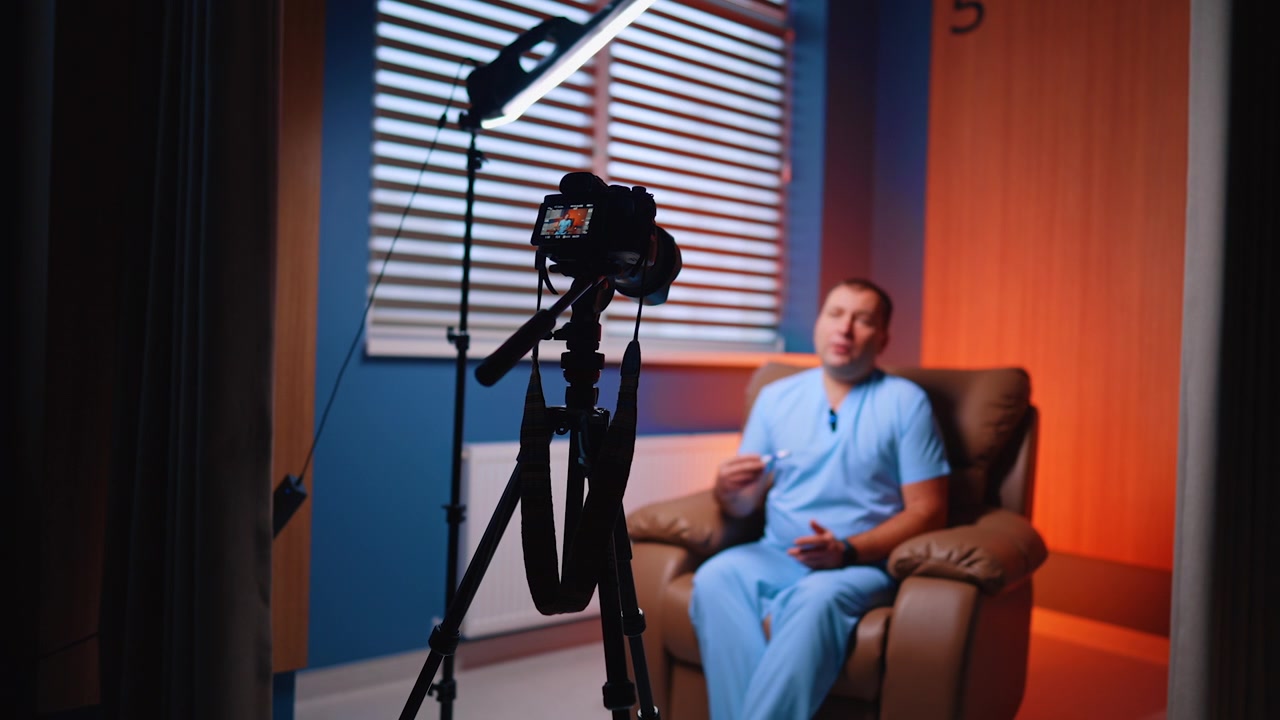 Statiefcamera filmt zittende man in oranje en blauwe kamer. Apparatuur op de voorgrond met een wazig figuur in ziekenhuiskleding, achteroverleunend in een leren fauteuil bij een raam.