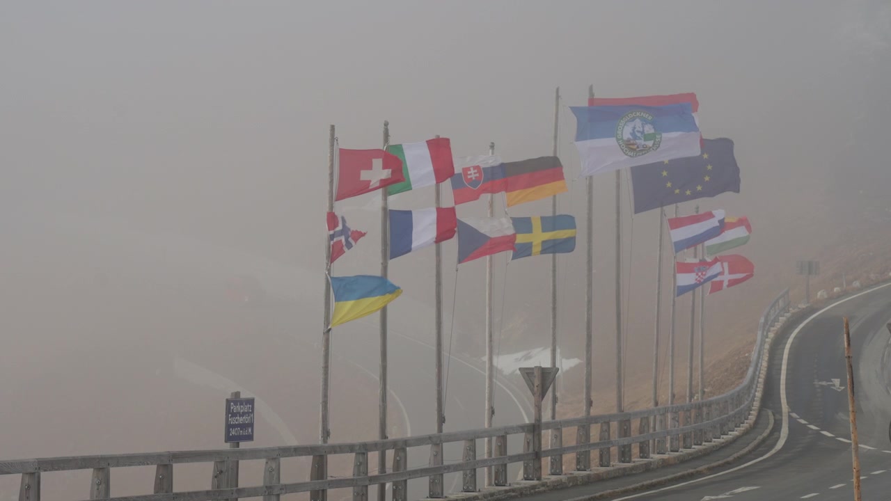Multiple flags of different countries flutter on tall poles as fog envelops the area, creating a soft, muted atmosphere along a curvy road.