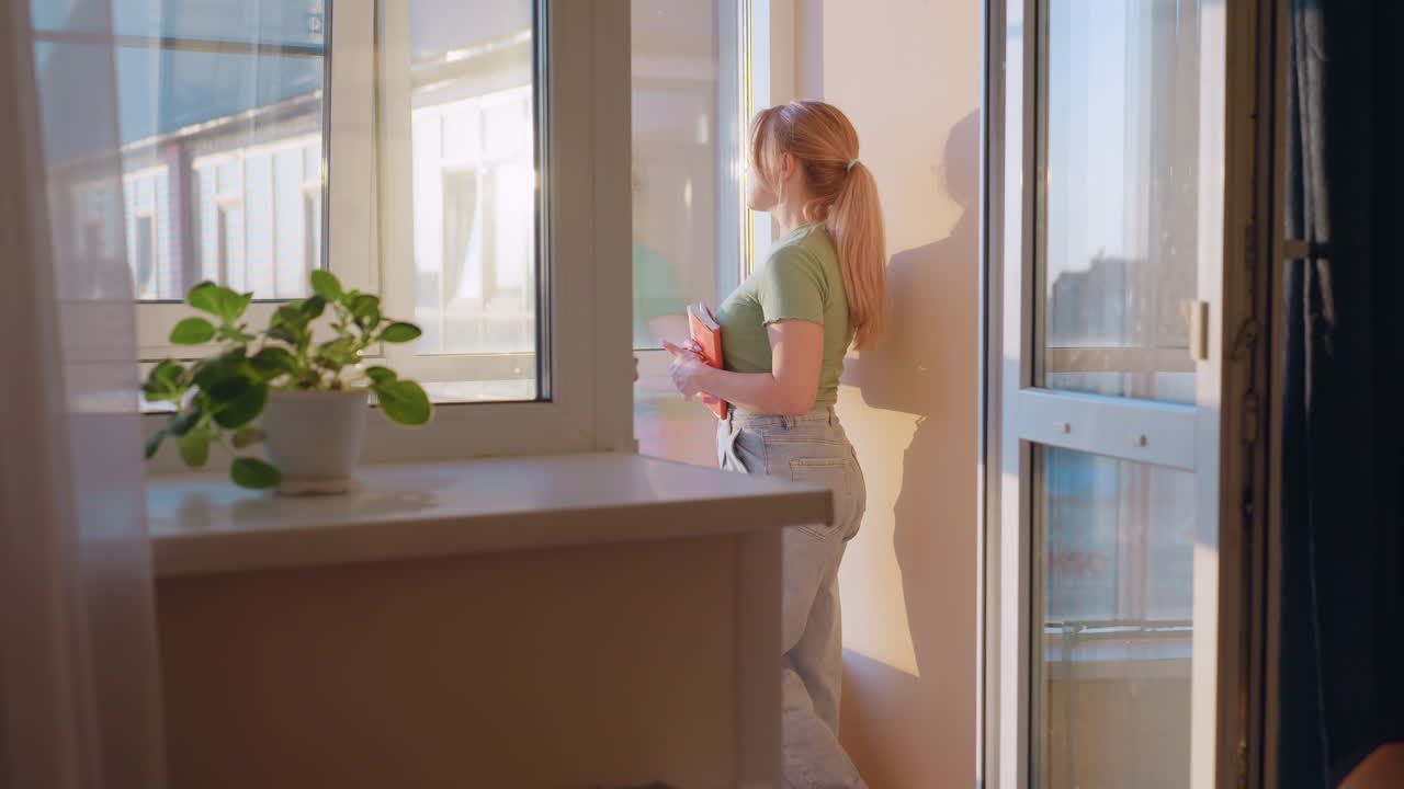 Woman holding book looking out thoughtfully through window during sunset, reflecting with calm and peaceful expression, sunlight casting warm glow indoors
