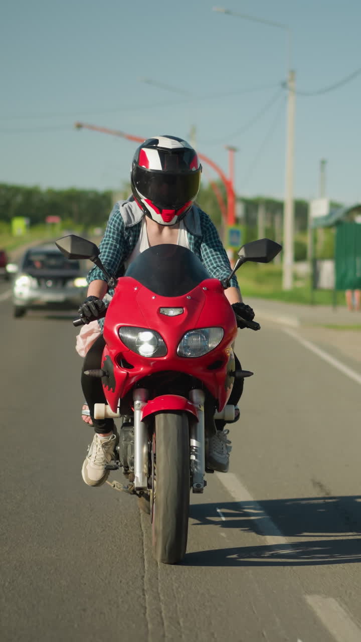 una motociclista femenina monta una motocicleta roja en un día soleado, navegando por una carretera rural con árboles, postes eléctricos y coches en el fondo, con coches viajando por la misma carretera