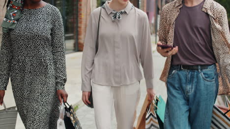 cheerful multiethnic friends walking with shopping bags and using smartphone