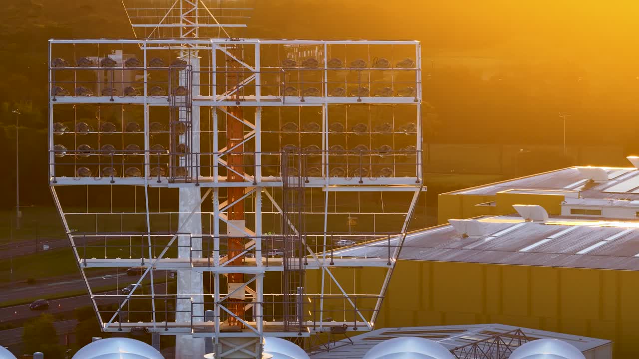Drone glides past large stadium floodlight tower at sunset, revealing sports venue, nearby buildings, and glowing sky with warm, golden lighting