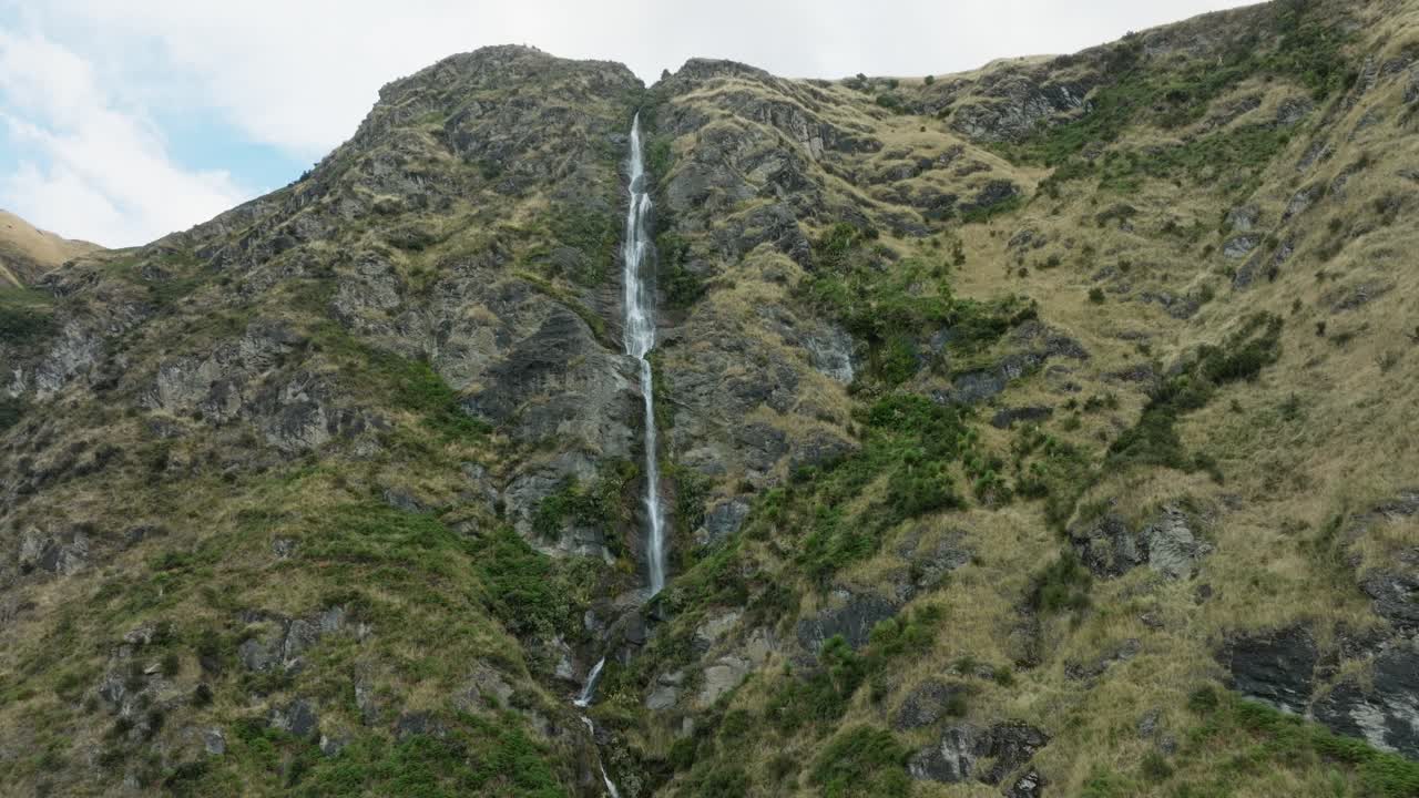 Scenic aerial view panning right to left of narrow, free flowing waterfall winding through rugged, rocky and mountainous landscape in the remote wilderness of New Zealand Aotearoa