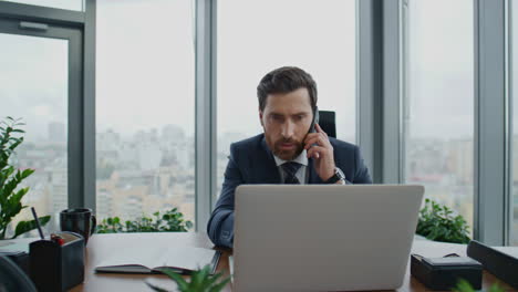 Director-Preocupado-Hablando-Por-Teléfono-Sentado-En-El-Escritorio-De-La-Oficina-Con-Un-Portátil-Moderno-De-Cerca.