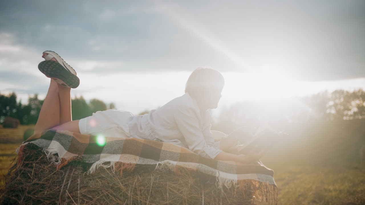 Woman lying on hay bale with checkered blanket, reading a book and flipping to a new page as glowing sunlight shines around her in serene open field
