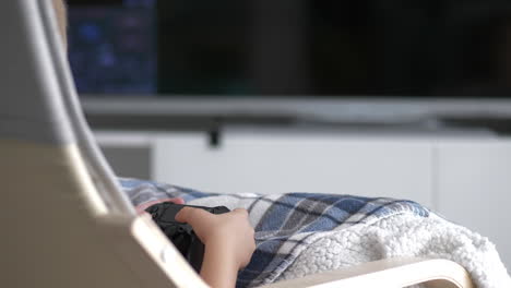 Unrecognized-caucasian-boy-holding-console-game-controller-and-pressing-buttons,-Sitting-on-chair,-indoors