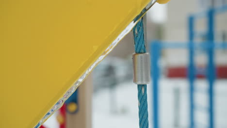 primer plano de un columpio amarillo y azul con detalles de helada en una cerradura de metal y una cuerda azul vibrante, el fondo sutilmente borroso con coloridos equipos de parque infantil