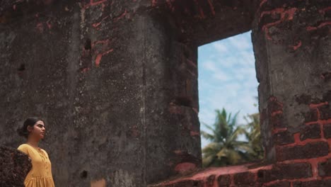 A-woman-in-a-yellow-dress-stands-by-an-old-stone-wall-with-a-window-opening-to-a-tropical-scene