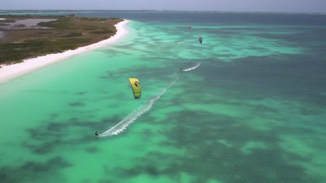 Kite-surfers-gliding-over-clear-turquoise-waters-near-a-sandy-beach,-aerial-view