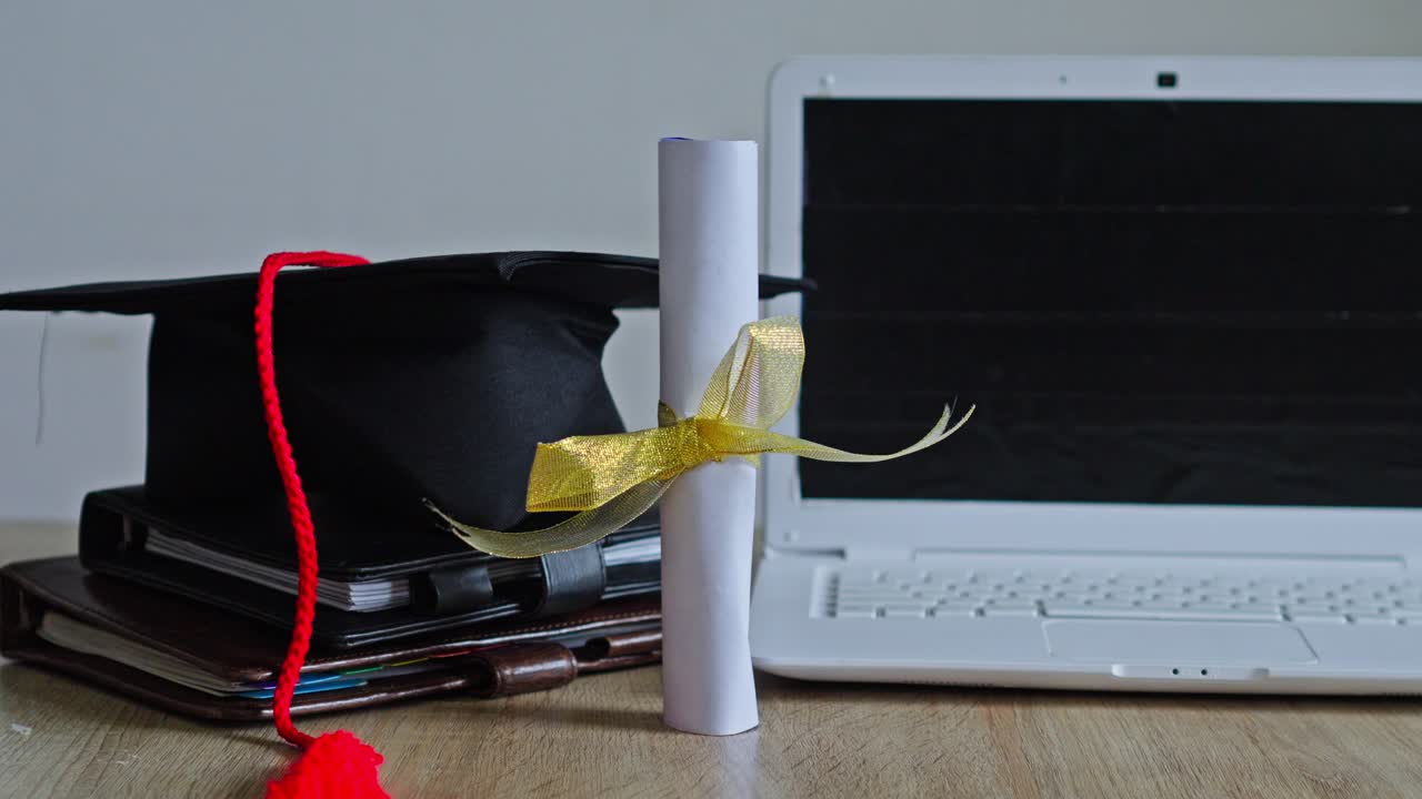 Mano che posiziona un diploma arrotolato con nastro dorato accanto a un tocco da laureato e un laptop bianco