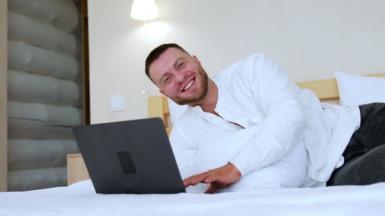 Bearded man in white shirt lies on the bed working on laptop. Portrait of a happy man laughing cheerfully.