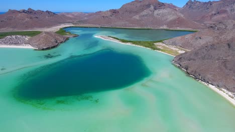 Slow-Forward-Aerial-of-Scenic-Blue-Hole,-Long-White-Sand-Beaches,-Green-Foliage-and-Desert-Mountains-in-Background