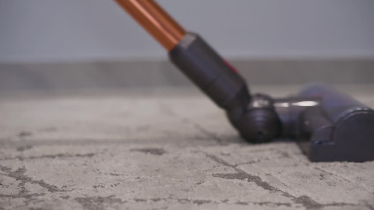 Low-angle close-up of a vacuum cleaner head moving across a carpeted floor inside a home. The video highlights household cleaning, modern appliances, and everyday domestic maintenance
