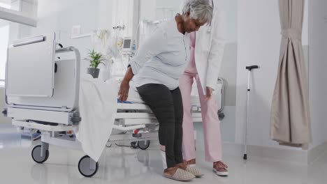 happy diverse female doctor helping senior female patient stand from bed, slow motion, copy space