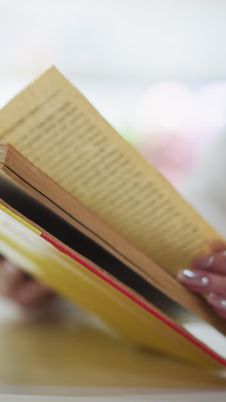 Close-up of hands flipping through pages of an open yellow book on a table, with a blurred background featuring a coffee cup, creating a serene and thoughtful atmosphere